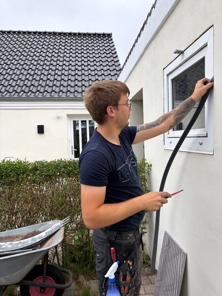 En mand med briller og tatoveringer arbejder med at installere eller justere et vindue på en hvid væg udendørs.