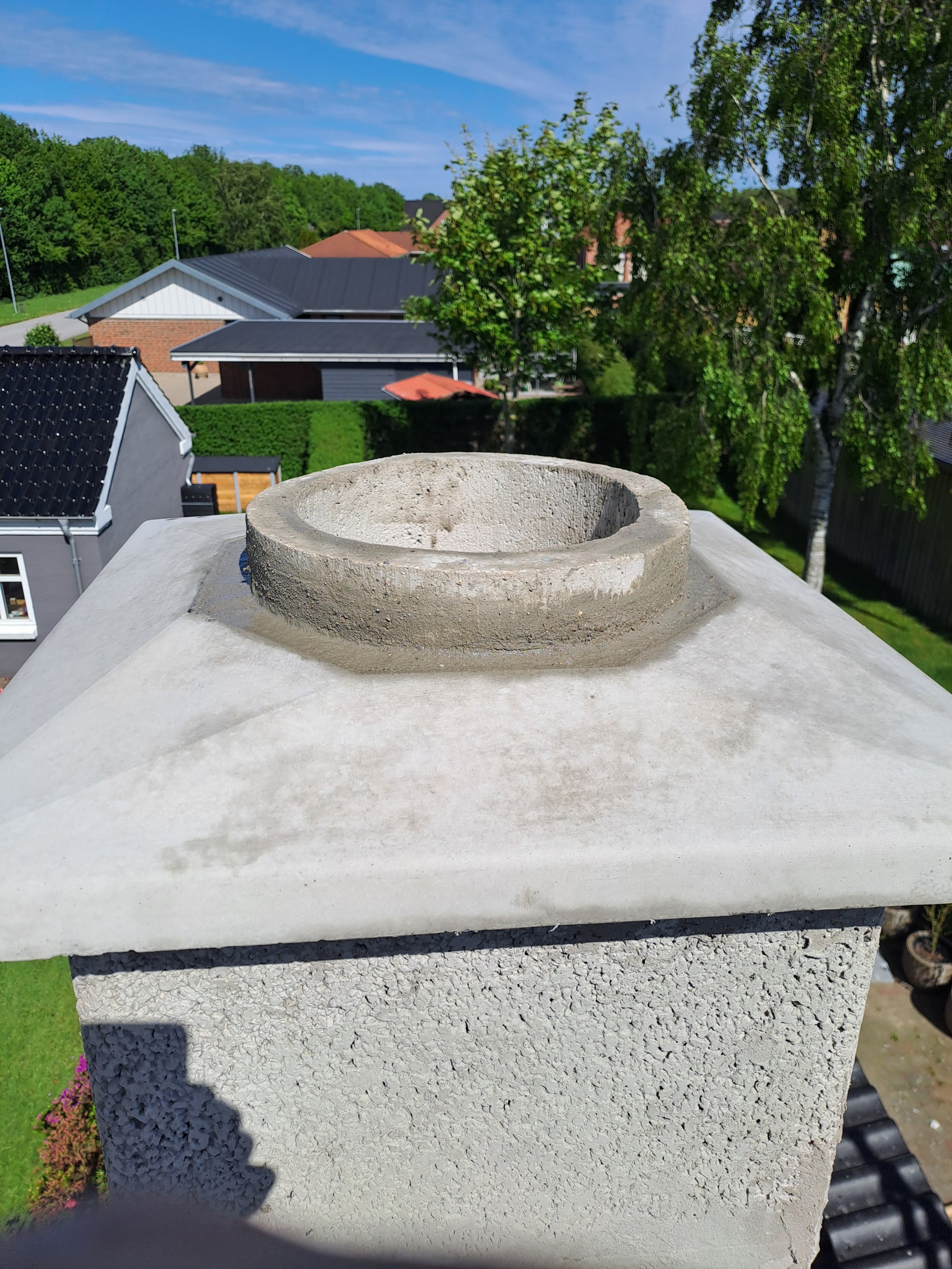 Billedet viser en støbt cementkappe eller sokkel, placeret på toppen af en hævet betonfundament i en udendørs boligområde med grønne træer og huse i baggrunden. Solskin og blå himmel er synlig.