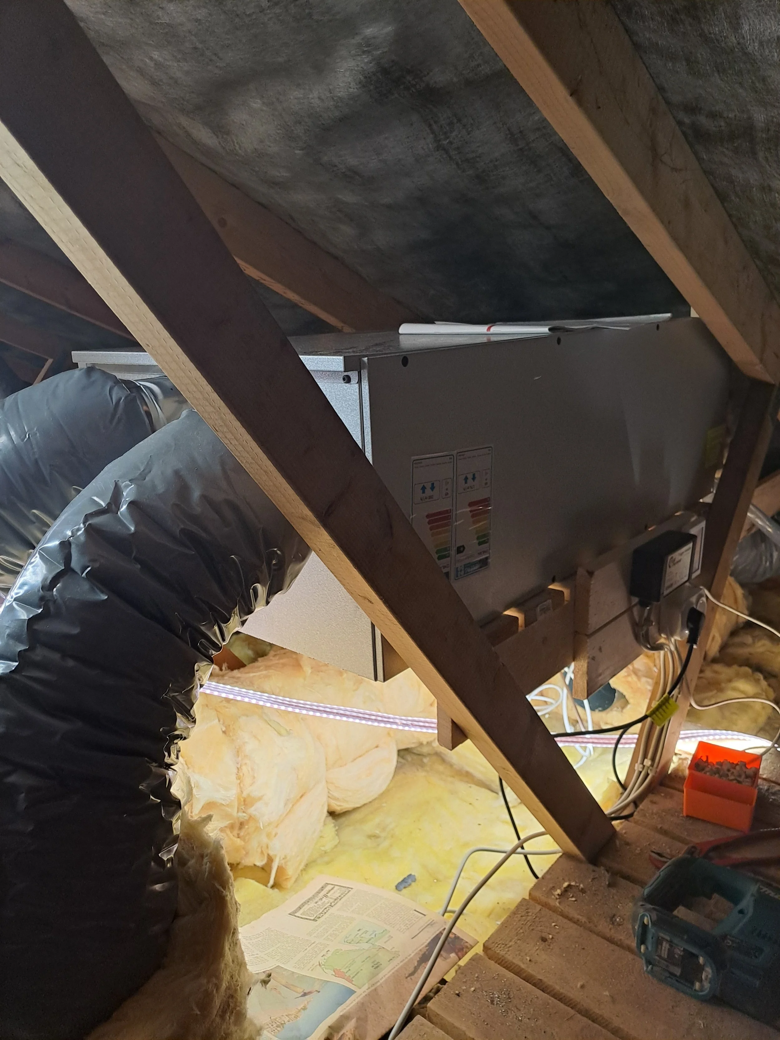 Udsigt af loft under konstruktion med udsatte træbjælker, isoleringsmateriale og værktøj, herunder en boremaskine, strømforsyningskabel og blade til udskæring.