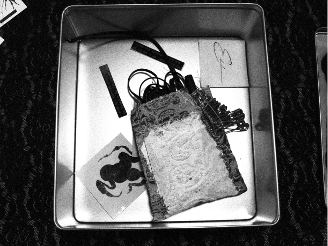 A metal box containing various office supplies, including rubber bands, paper clips, a printed image of a dog, and cut-out paper pieces.