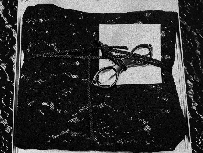 A black lace fabric wrapped around a white box, tied with a black cord and decorated with a metallic ring and some keys.
