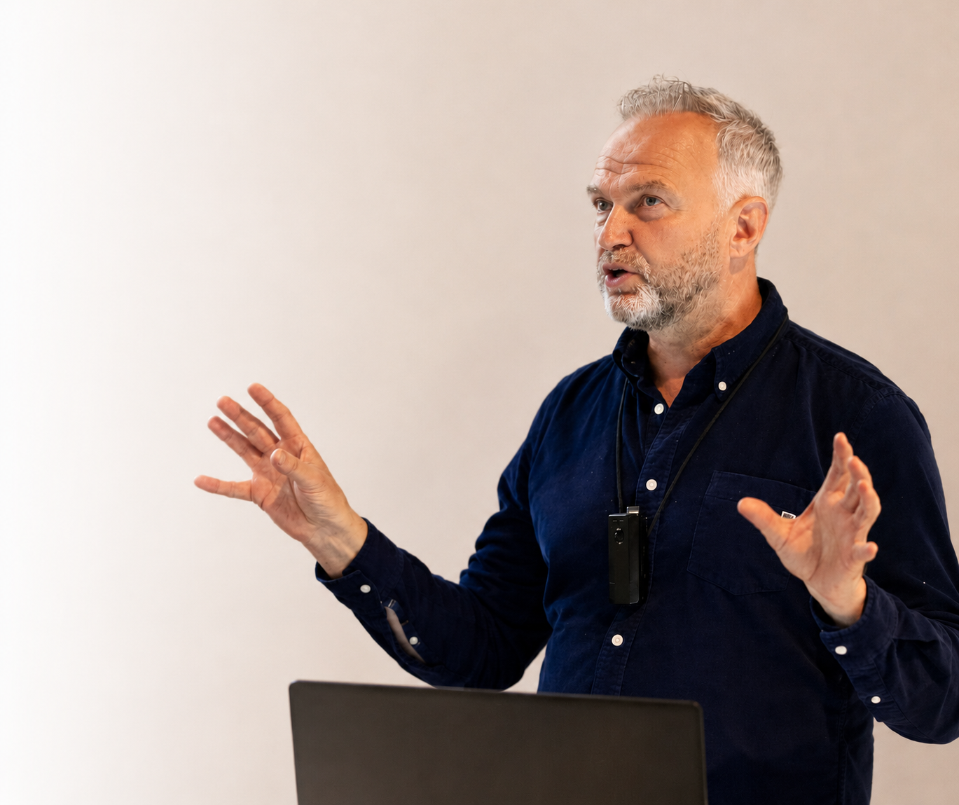 A middle-aged man with grey hair and beard, wearing a navy blue shirt, stands in front of a plain white wall, gesturing with his hands while speaking.