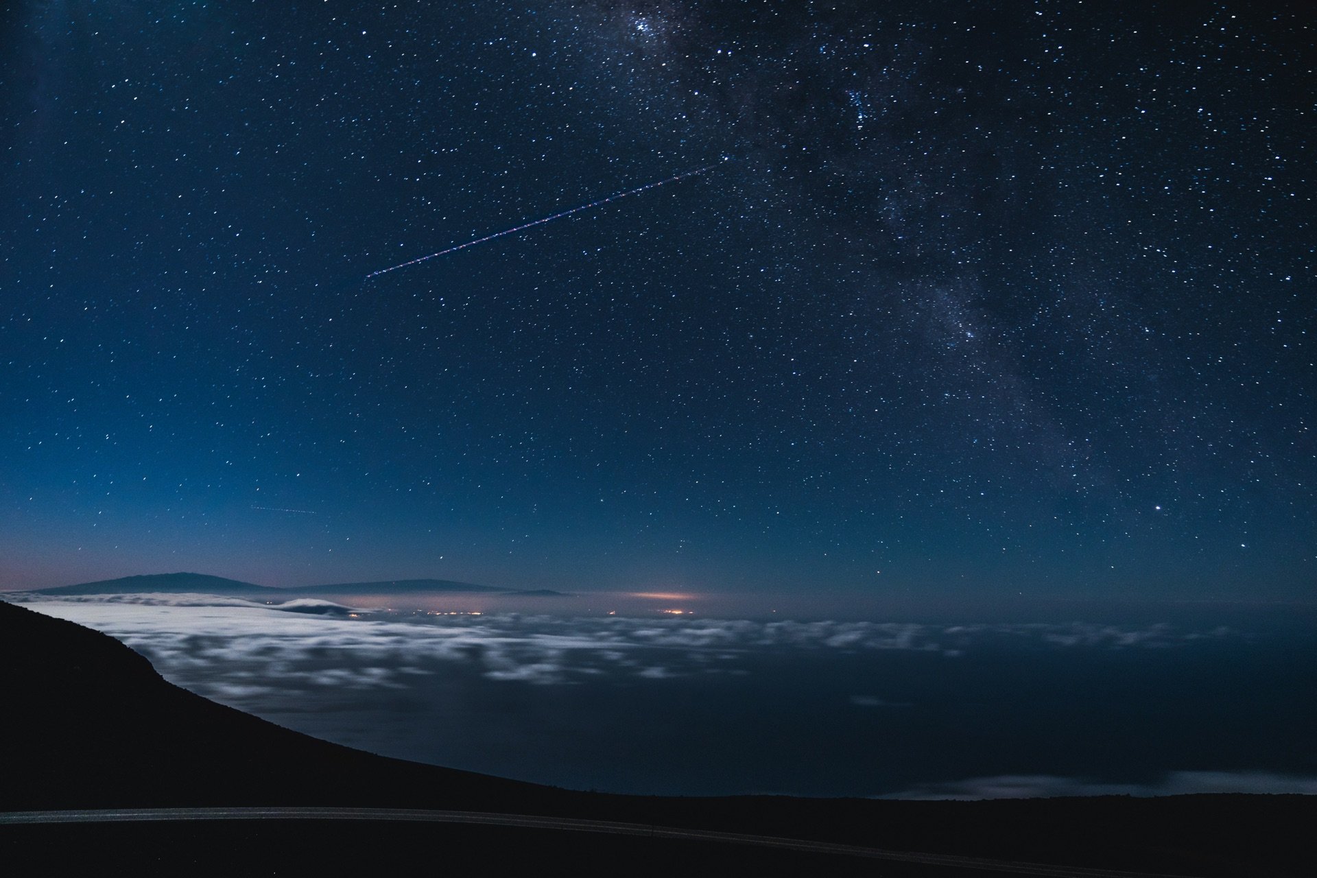 Night sky filled with stars and a visible Milky Way galaxy, with a shooting star streaking across, over a hilly landscape with clouds below.
