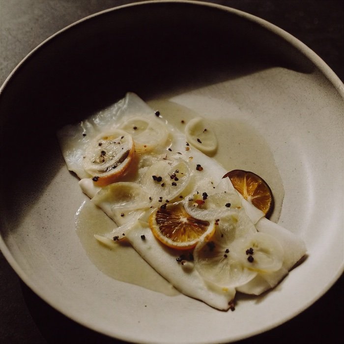 A white fish fillet topped with lemon slices, black pepper, and some sauce, served on a beige and gray ceramic plate.
