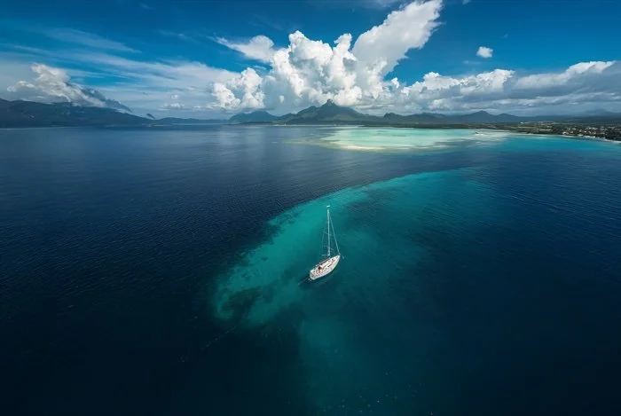 An aerial view of a sailboat anchored near a coral reef in a turquoise ocean, with a distant island, mountains, and cloudy sky in the background.