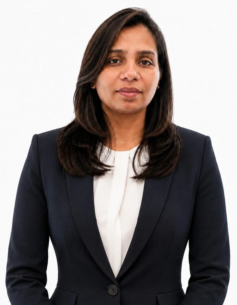 A woman with dark brown hair, wearing a black blazer over a white blouse, standing against a plain white background.