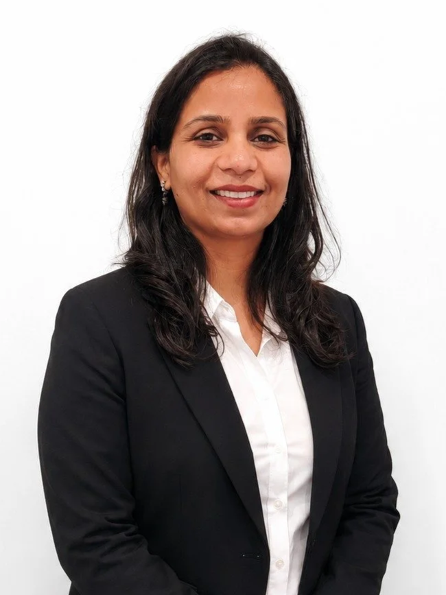A woman with dark brown hair, wearing a black blazer over a white blouse, standing against a plain white background.