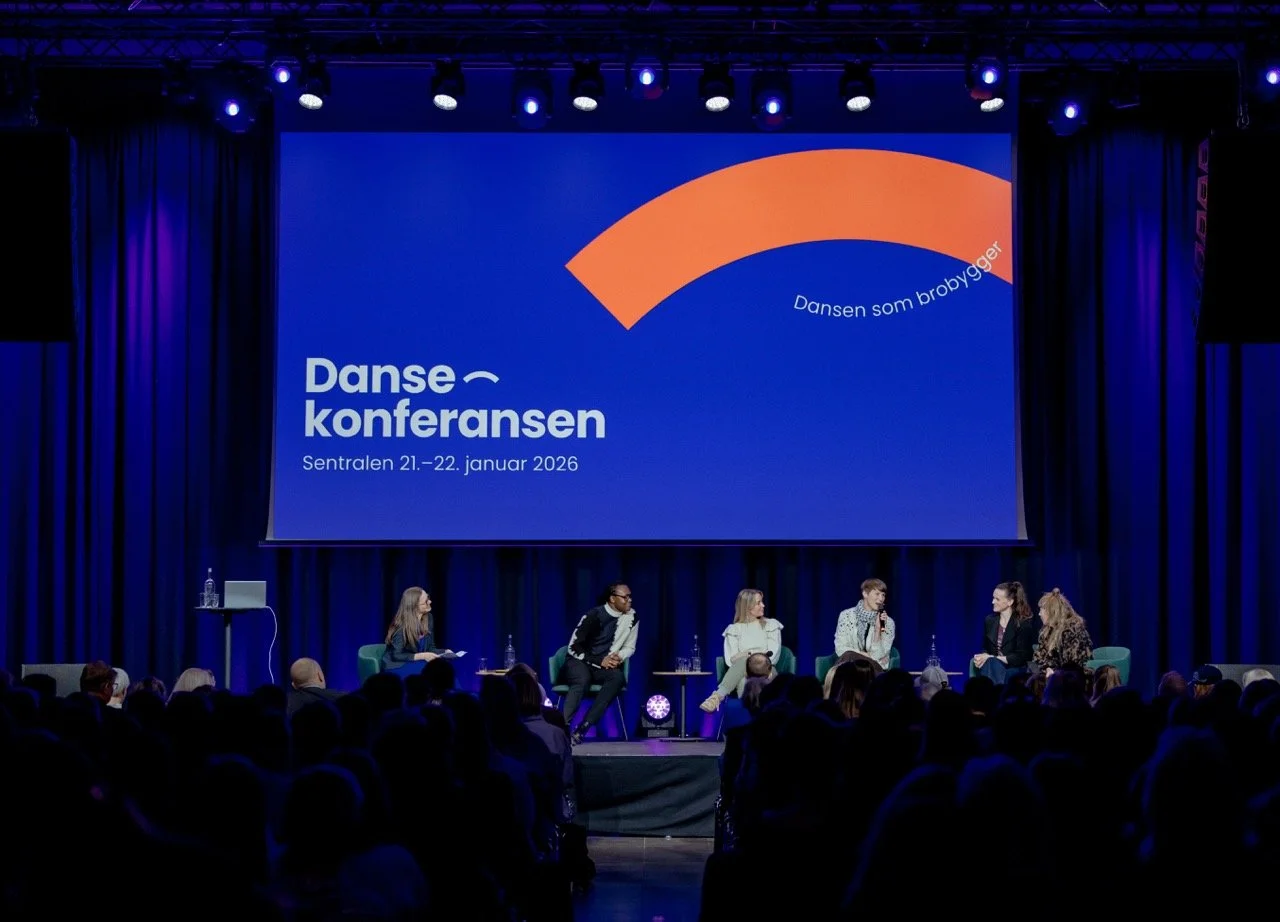 Flerfarget scene fra en konferans med flere personer på scenen, en stor skjerm som viser tekst om dansekonferansen 2026, og et fullt publikum.