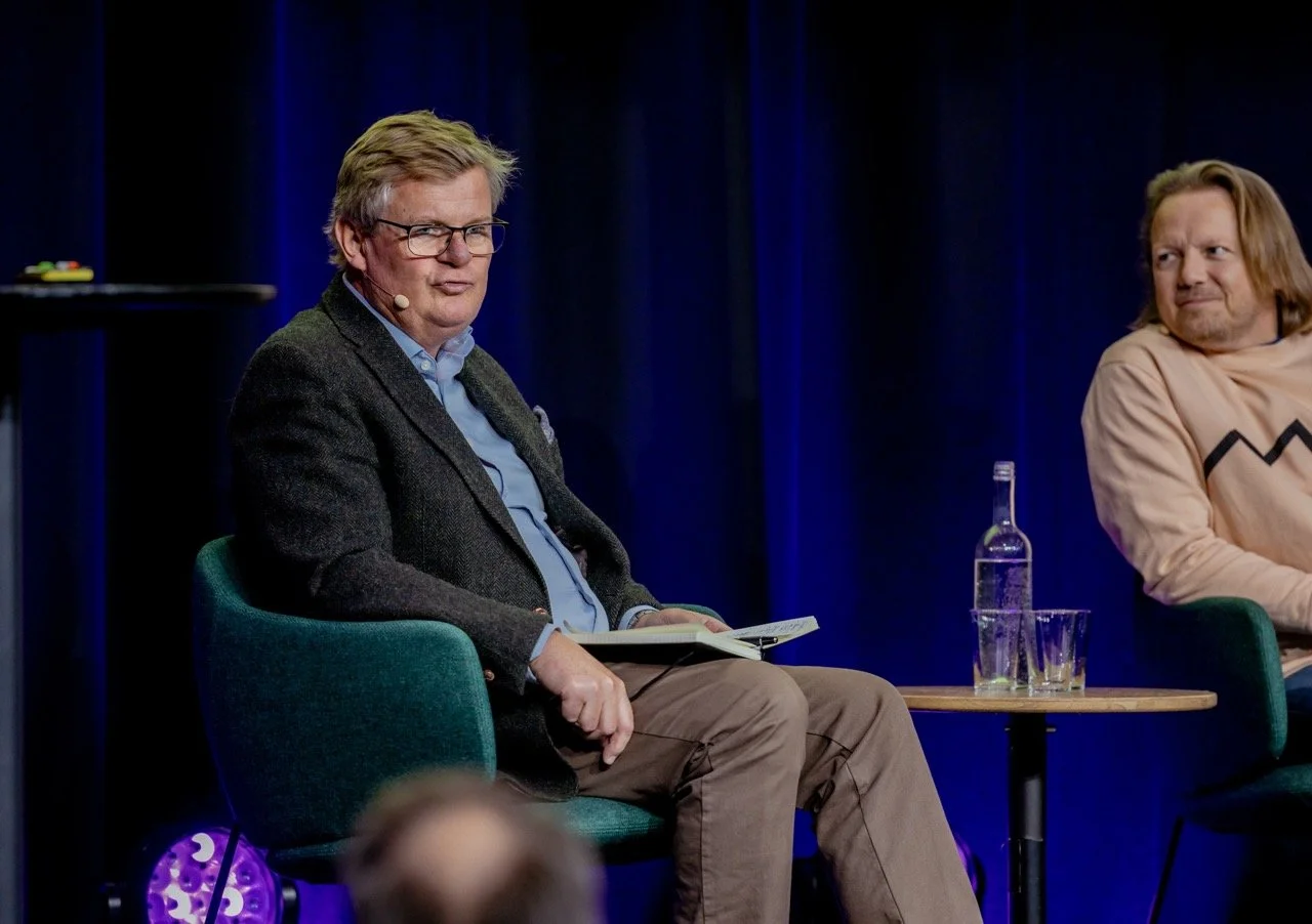 To menn sitter på en scene under en diskusjon eller panel. Den ene mannen har på seg briller og en mørk jakke, den andre har langt, rødbrunt hår og en beige genser. Det er en liten bord med en vannflaske og to glass foran dem.