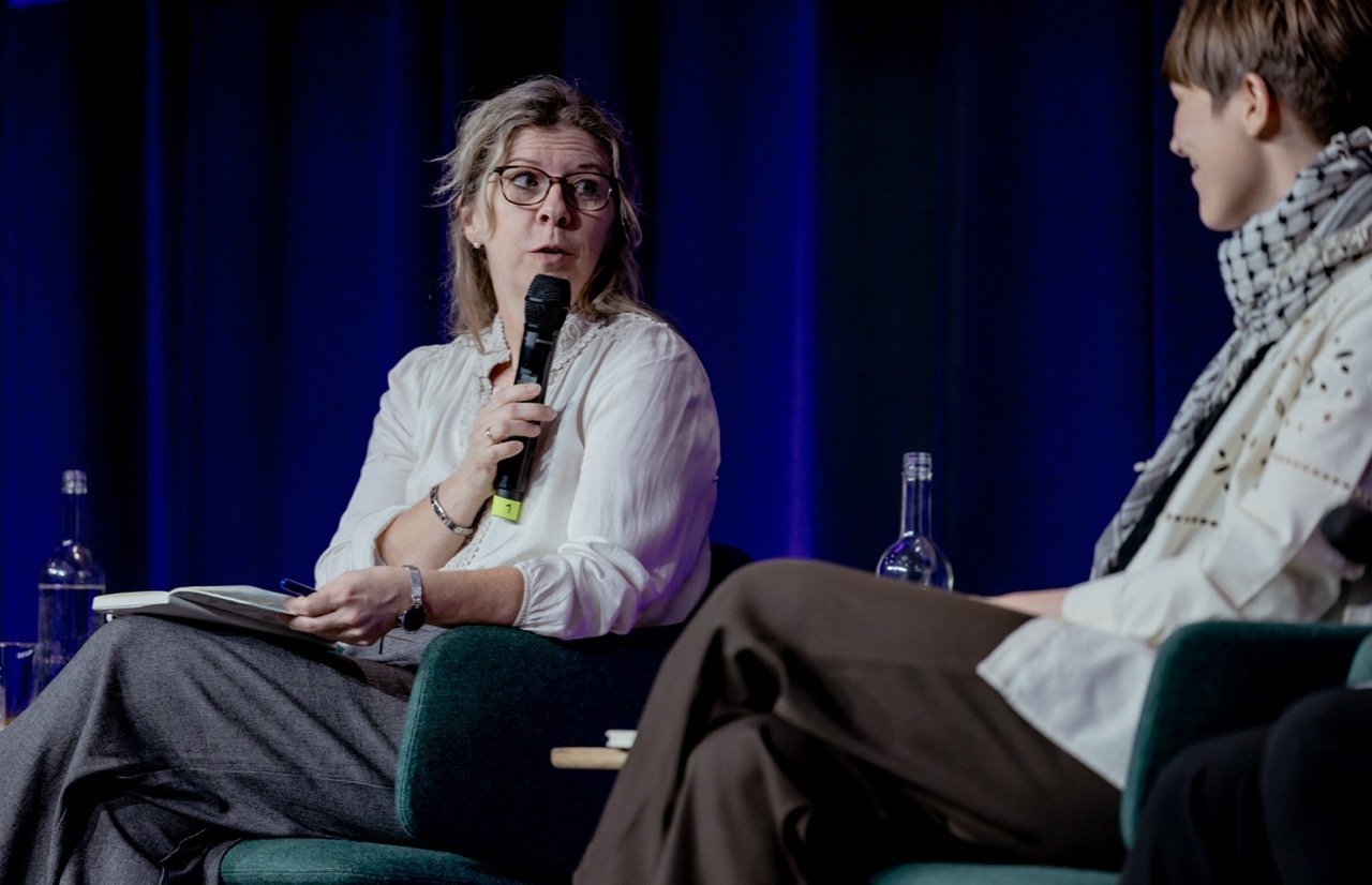 To personer i en paneldebatt, en kvinne med briller og en mikrofon, og en yngre mann, sitter i komfortable stoler på en scene foran en blå gardin.
