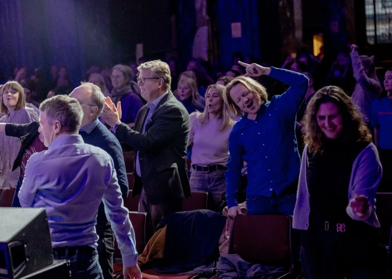 Folk som står og danser eller syns å ha det gøy i en offentlig setting, sannsynligvis et arrangement eller konsert, med mørk innendørs bakgrunn.