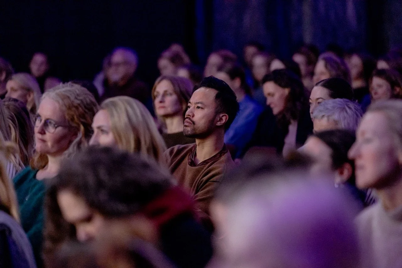 Et publikum som sitter i et mørkt auditorium under en presentasjon eller forelesning, med fokus på en mann i midten som ser ut til å følge med oppmerksomt.
