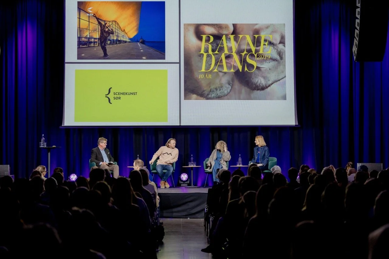 En paneldebatt med fire personer på scenen foran et stort lerret med bilder og tekst, i en teatersal med publikum i forgrunnen.