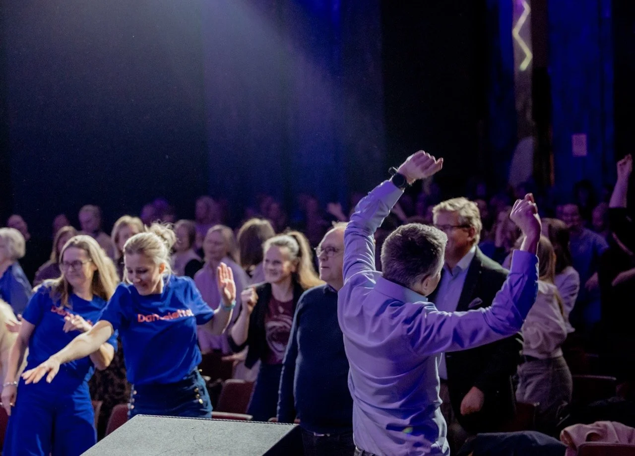 Mennesker danser og har det gøy på en konsert eller et arrangement med mørk bakgrunn og scenelys.