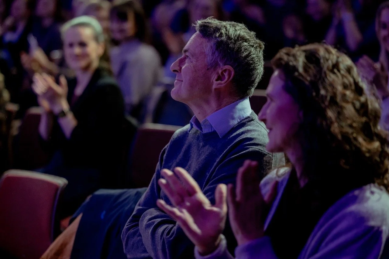 En gruppe mennesker som ser ut til å være på en konsert eller forestilling, alle klapper og smiler, med fokus på en mann i midten som har grått hår og kledd i en blå skjorte og grå genser.