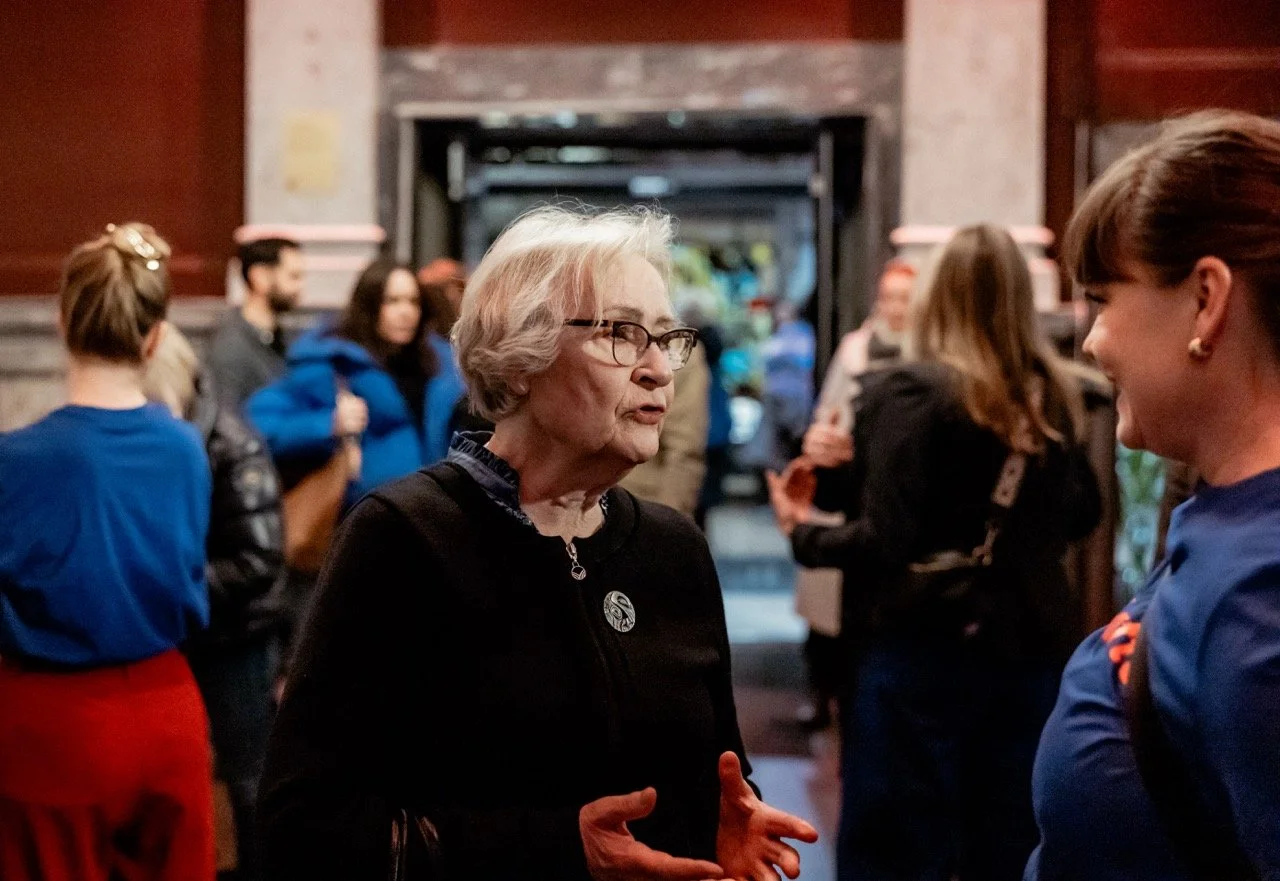 To eldre kvinner snakker med hverandre i en gruppe innendørs, med flere mennesker i bakgrunnen.