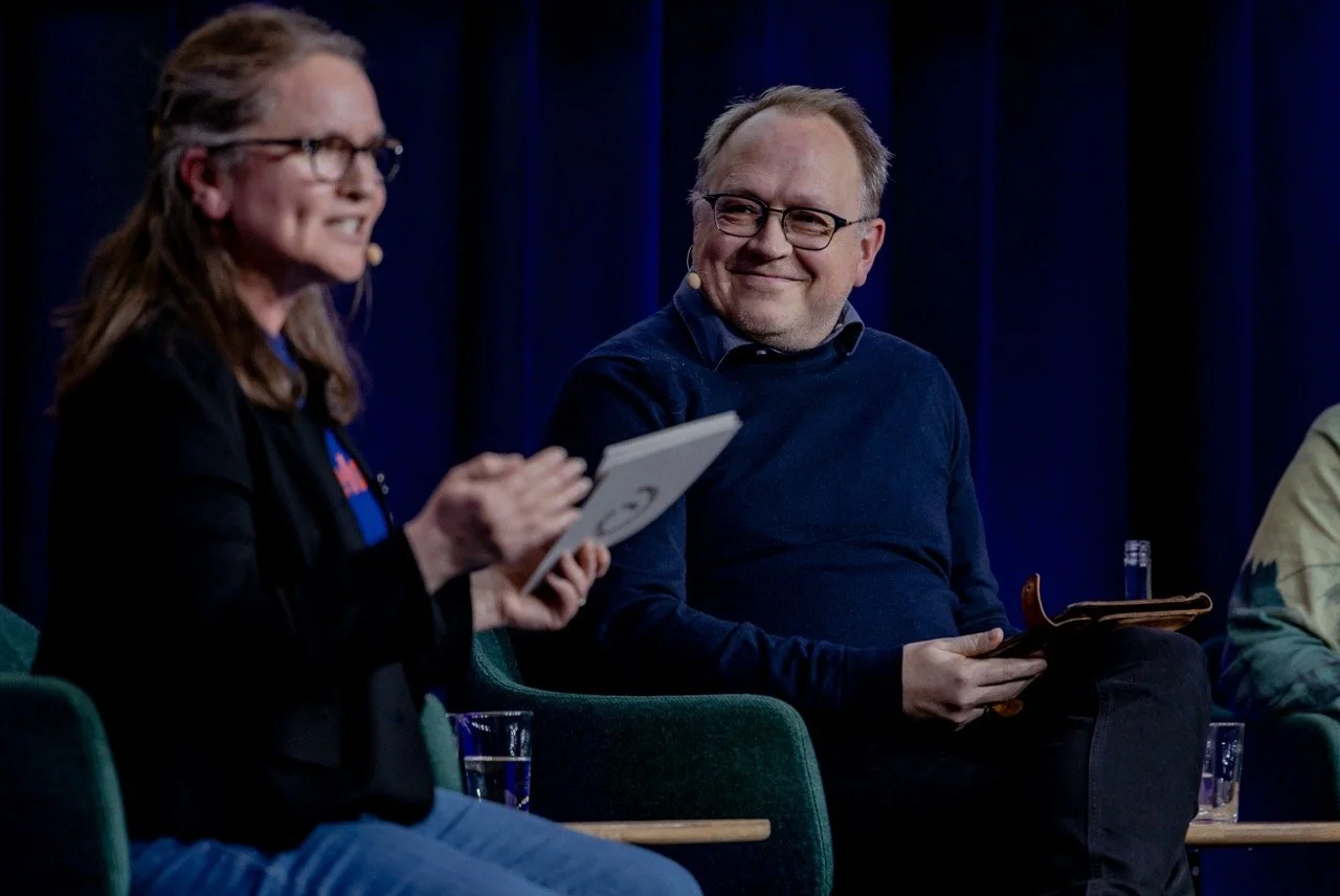 To personer som sitter i en paneldebatt, smilende og snakker, med mørk bakgrunn og grønne stoler.