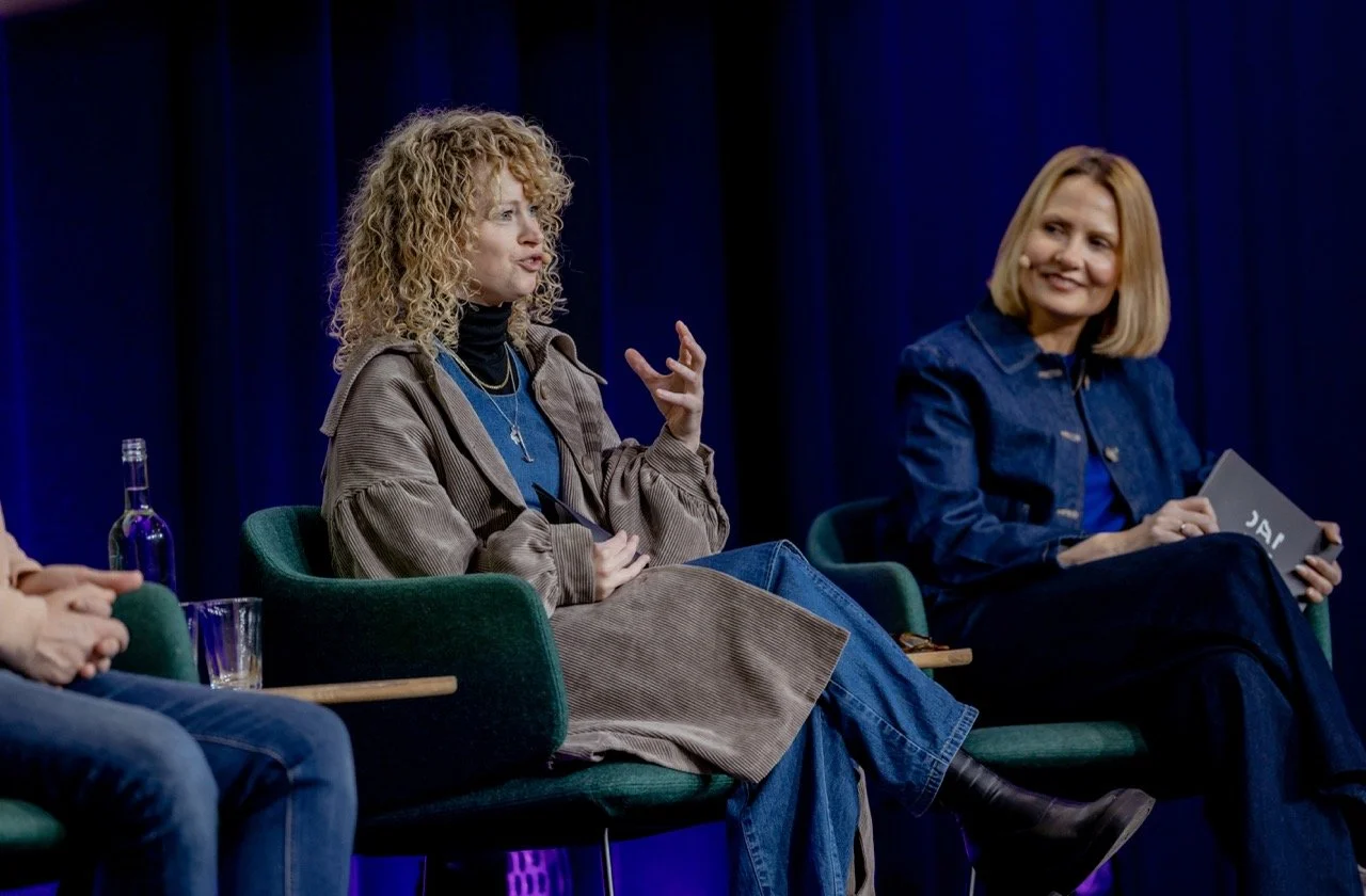 To kvinner som snakker på en paneldebatt, sittende i grønne stoler foran en mørk blå backdrop. Den kvinnen til venstre har krøllete, blond hår og snakker, mens kvinnen til høyre har rett, blond hår og smiler.