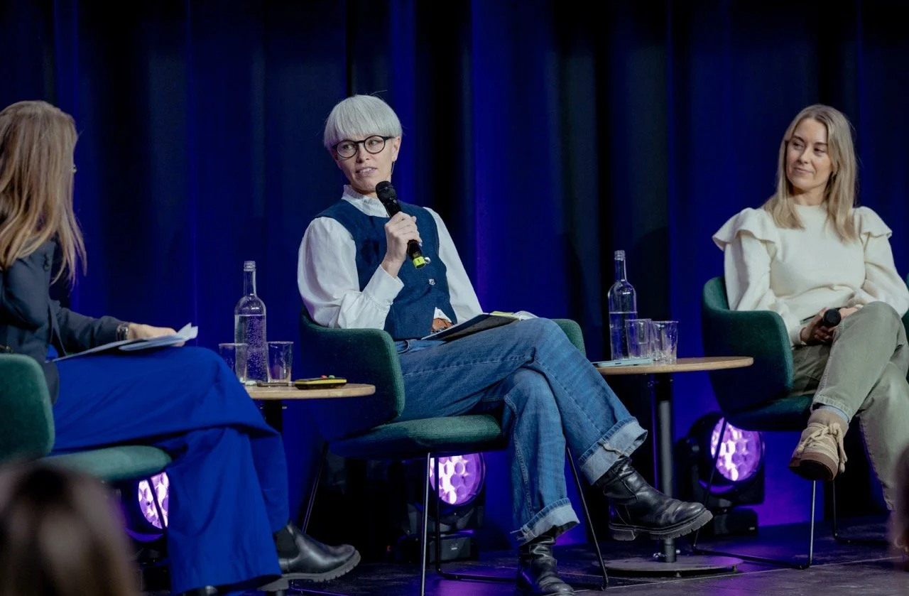 Tre kvinner på en scene under en paneldiskusjon, med mikrofoner, vannflasker og glass på bordet foran dem, bakgrunnen er mørk med blå belysning.