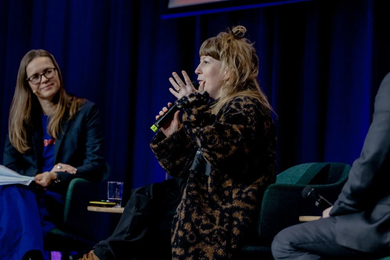 En kvinne snakker inn i en mikrofon under en paneldebatt, mens en annen kvinne ser på og smiler.