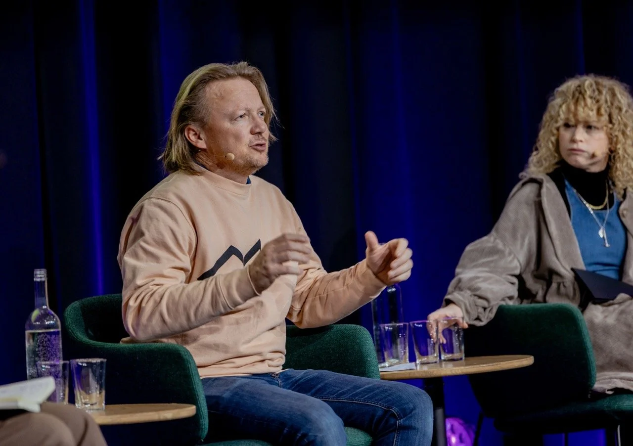 To personer som sitter på stoler på en scene, en mann snakker og en kvinne lytter.
