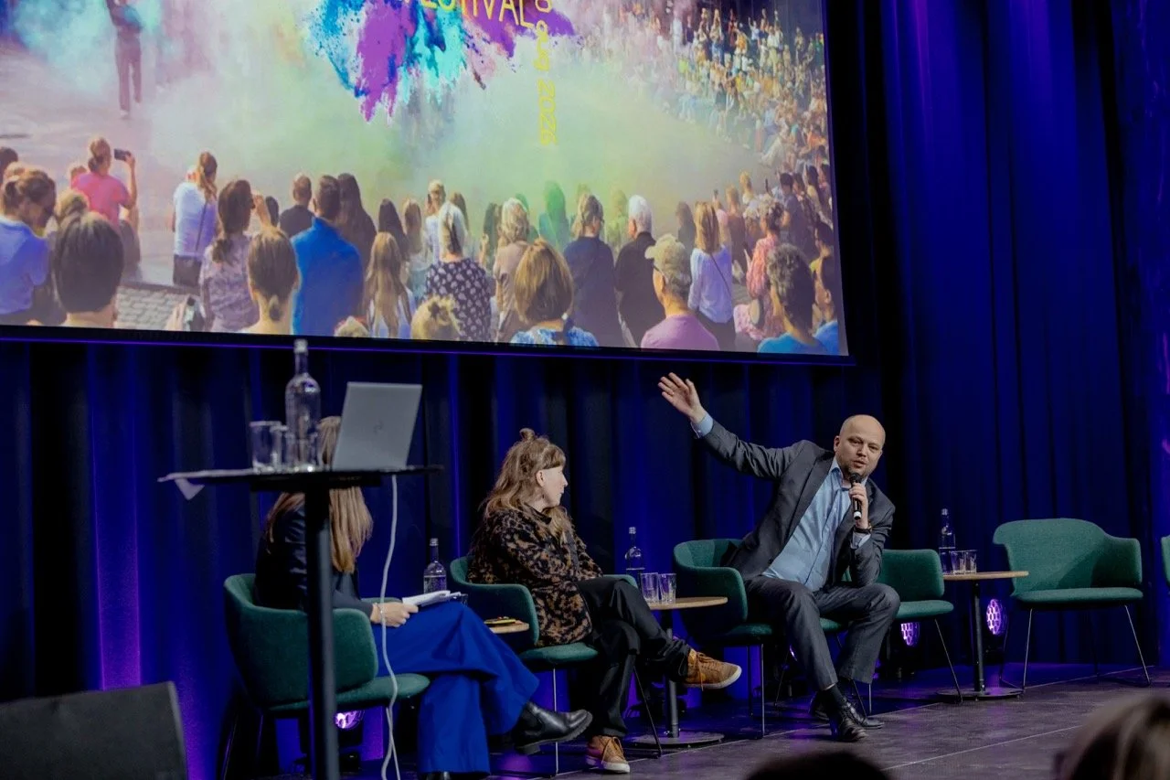 En paneldiskusjon med tre personer på scenen foran en stor skjerm som viser et bilde av en folkefest med mange mennesker og fargepulver, på en scene med blå gardiner og publikummer foran.