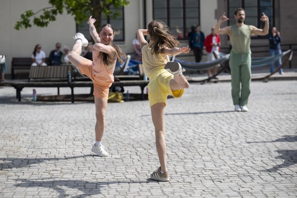 To unge jenter danser med en mann i en offentlig plass, med andre mennesker i bakgrunnen.