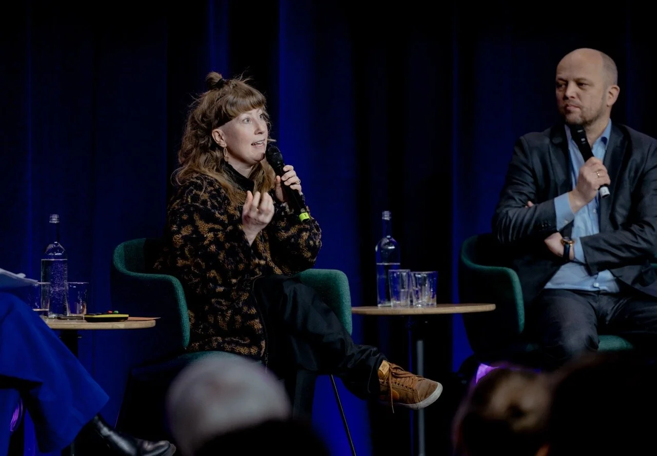 En kvinne snakker i mikrofon under en paneldiskusjon, mens en mann sitter ved siden av henne. Begge holder mikrofoner og er på scenen med mørkeblå bakgrunn.