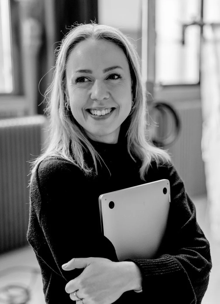 Kvinne med blond hår smilende, holder en laptop, i et rom med vinduer og radiator.