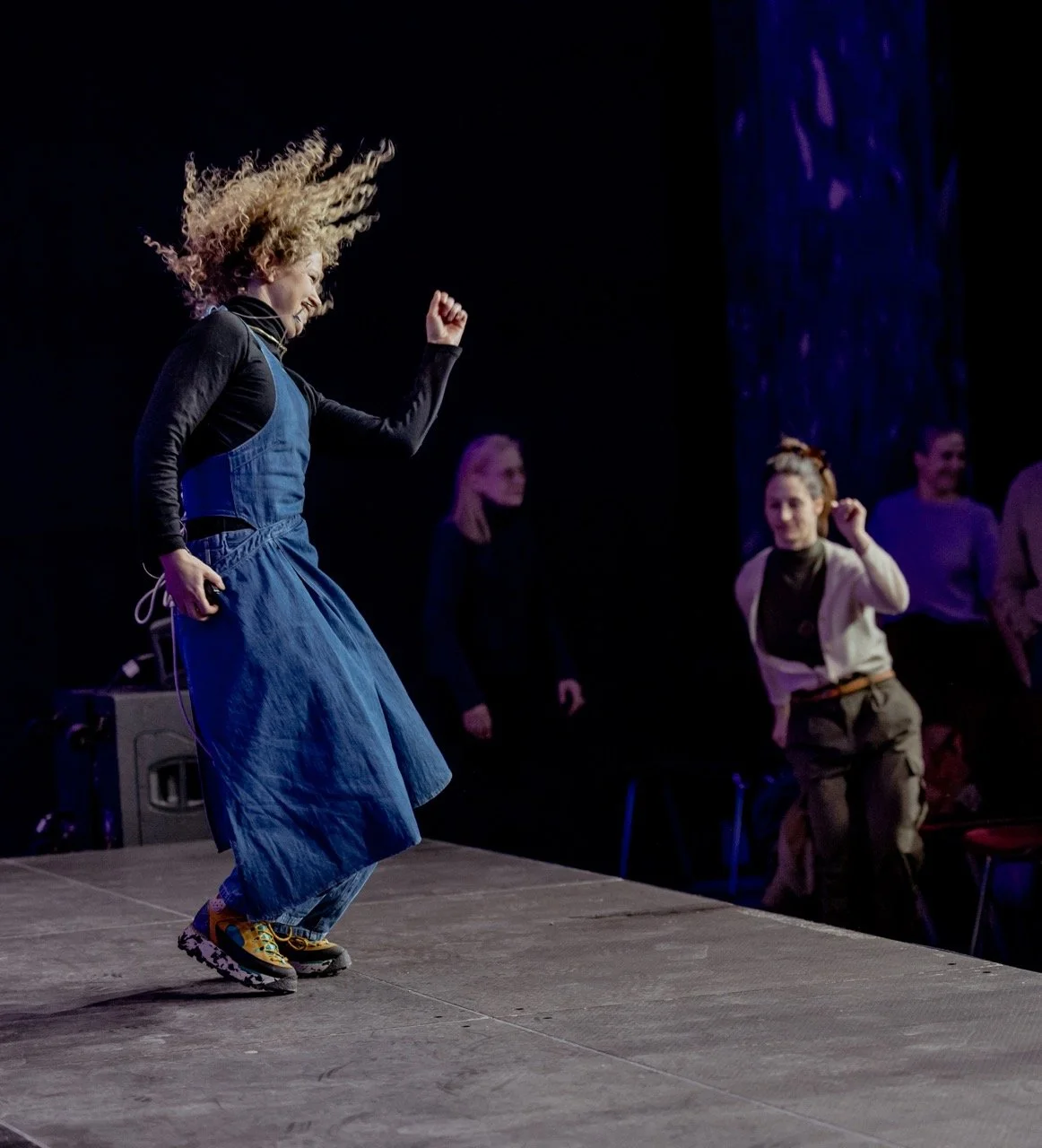 En kvinne danser på en scene med andre personer i bakgrunnen, hun har krøllete blondt hår og er iført en svart langermet topp, blå kjole og fargerike sneakers.