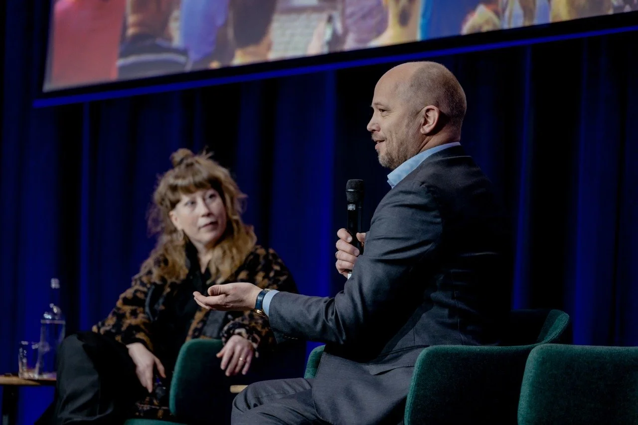 To personer snakker under en paneldebatt på en scene med blå bakgrunn. En mann holder mikrofon og snakker, en kvinne ser på ham.