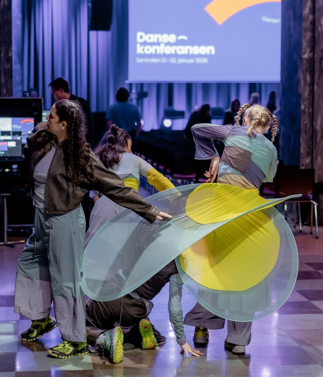 Dansegruppe som opptrer med bøyede kropper, noen som står og noen som ligger på gulvet på en scene, med stor skjerm i bakgrunnen som viser teksten "Danses konferansen".