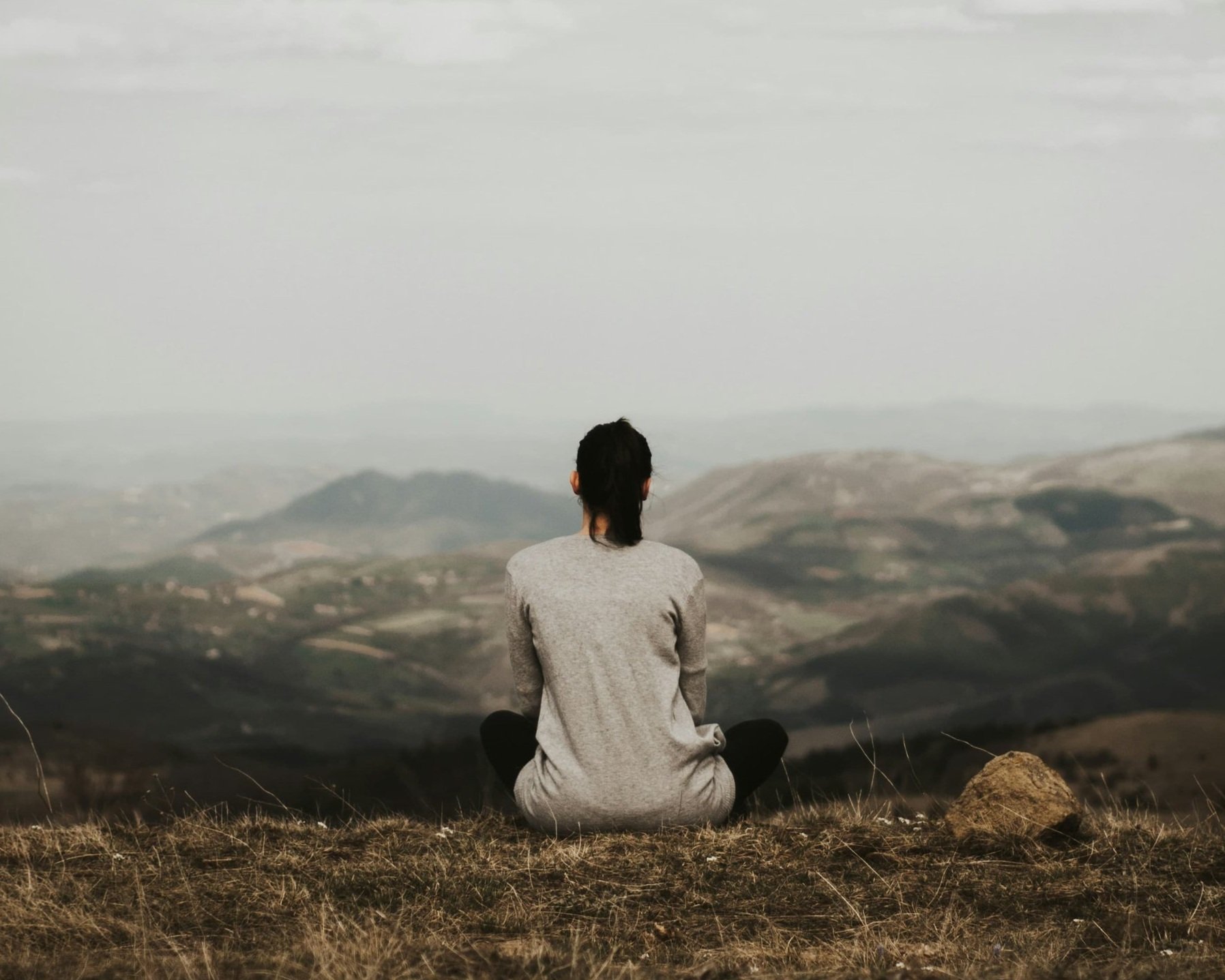 Person sitting cross-legged on a hilltop, overlooking a landscape of hills and valleys with a cloudy sky overhead.