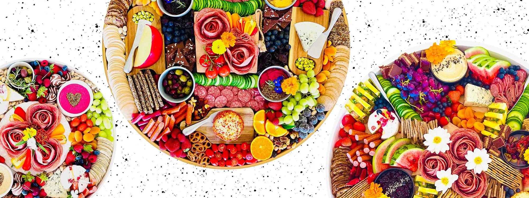 Colorful charcuterie boards featuring a variety of fruits, vegetables, cheese, chocolates, crackers, and decorative flowers on a white speckled background.