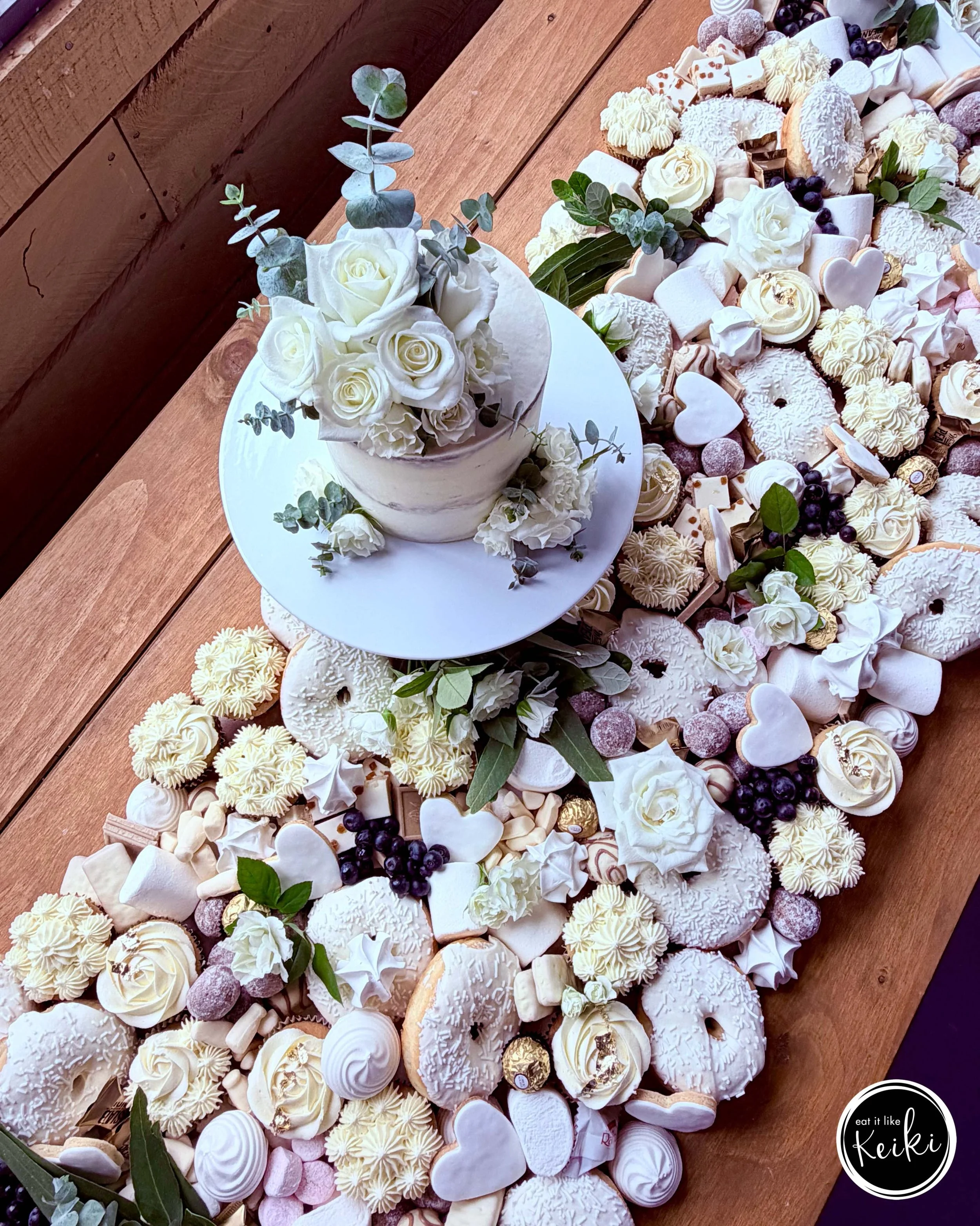 A large assortment of white and cream-colored desserts, including cupcakes, cookies, meringues, and candies, arranged on a wooden table with green leaves for decoration. There is a small circular logo in the bottom right corner that says 'eat it like Keiki.'