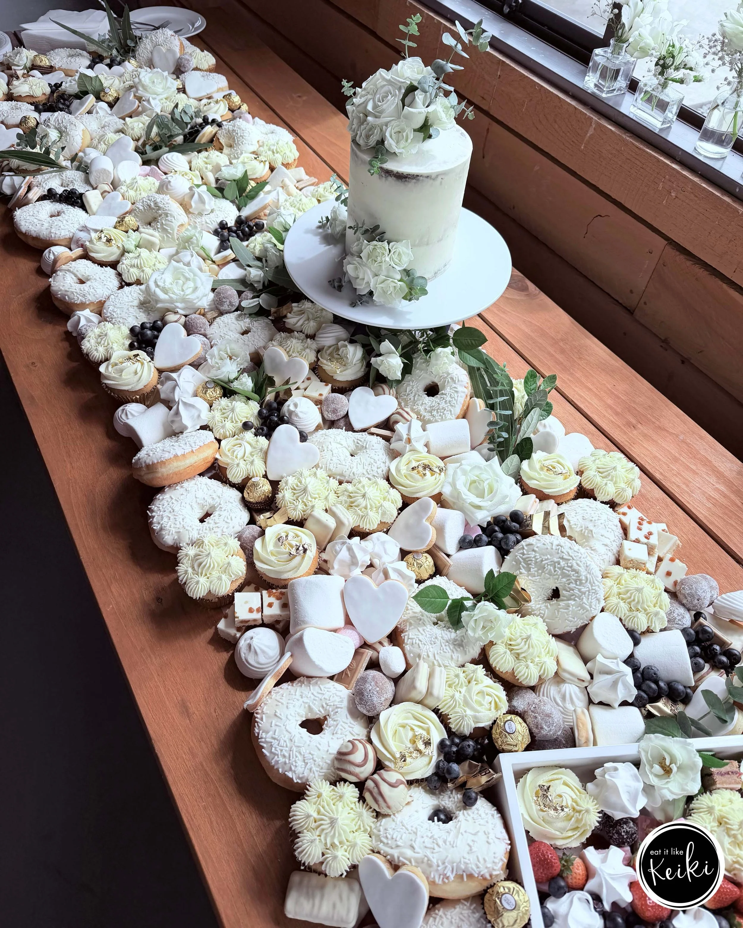 A large assortment of decorated cookies and sweets spread across a wooden table, with a white cake decorated with white roses and eucalyptus leaves in the center. Small vases with white flowers are placed on the windowsill.