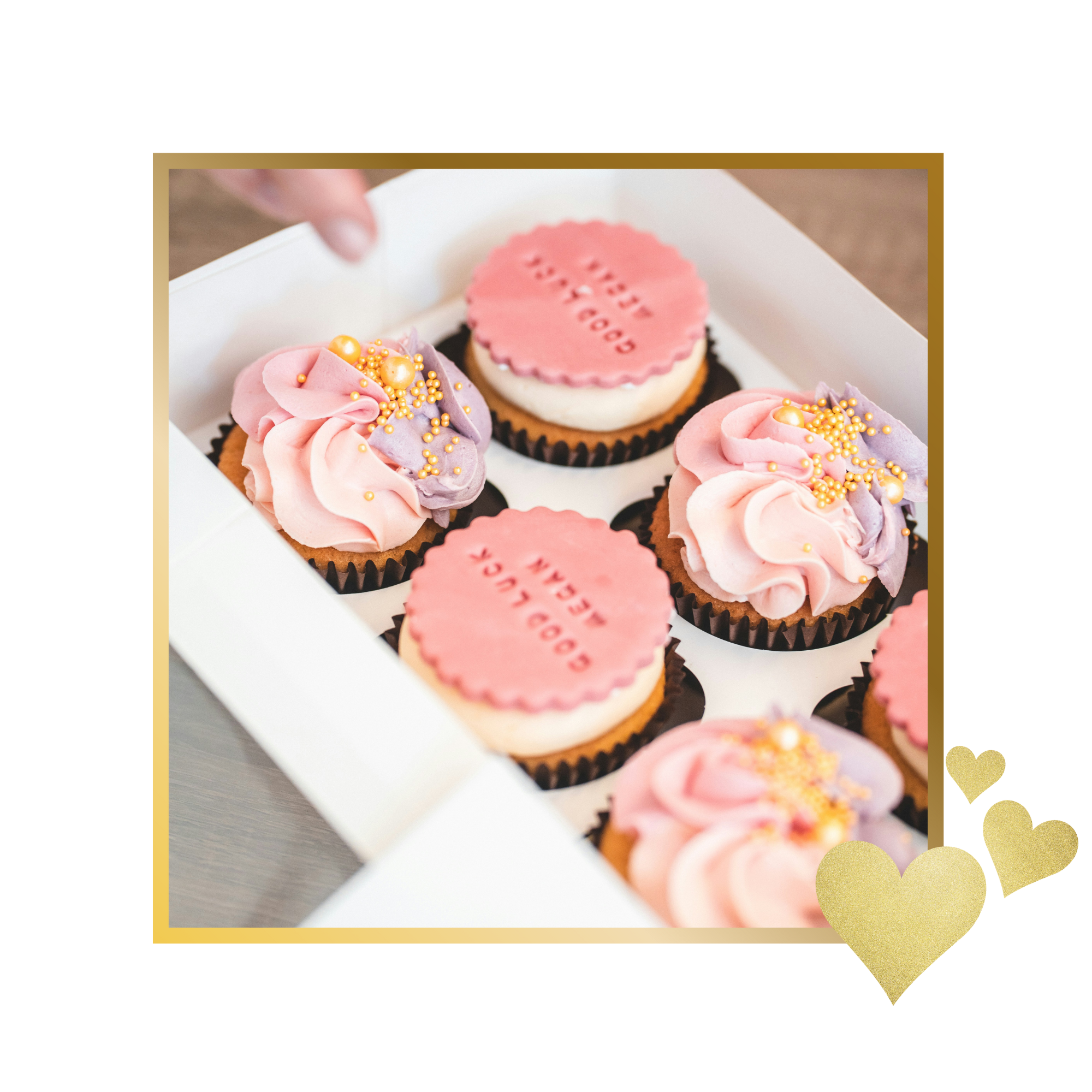 Box of decorated cupcakes, some with pink and purple icing swirl topped with gold sprinkles, and others with pink fondant rounds that read "Good Luck" and "Dream".