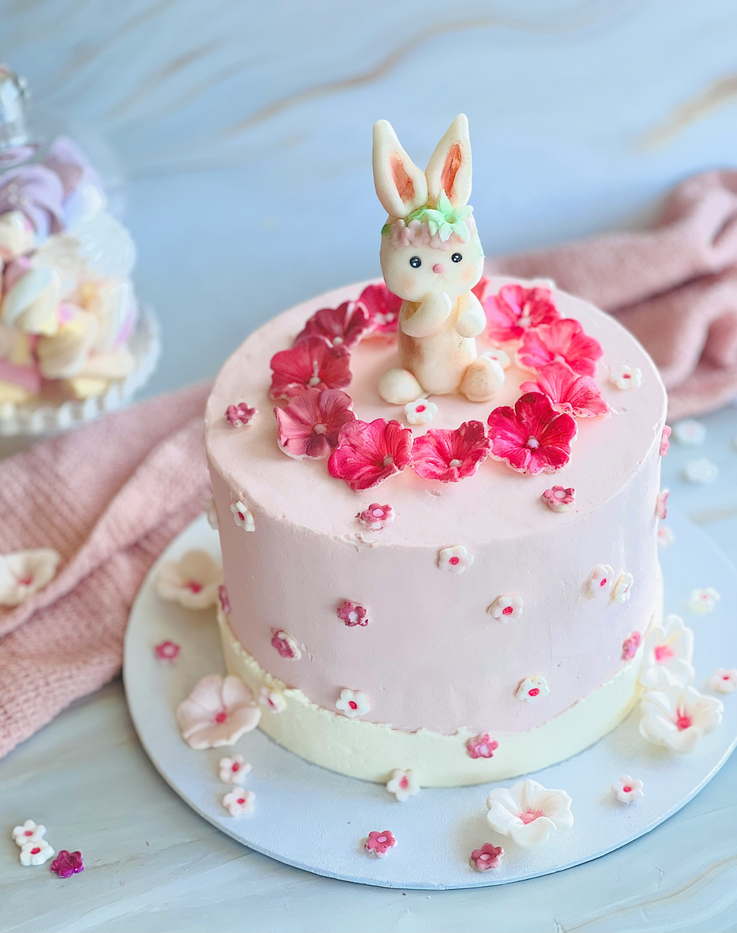 A pink birthday cake decorated with small white and pink flowers, topped with a fondant bunny figurine with big ears and a floral crown, surrounded by scattered flower petals.