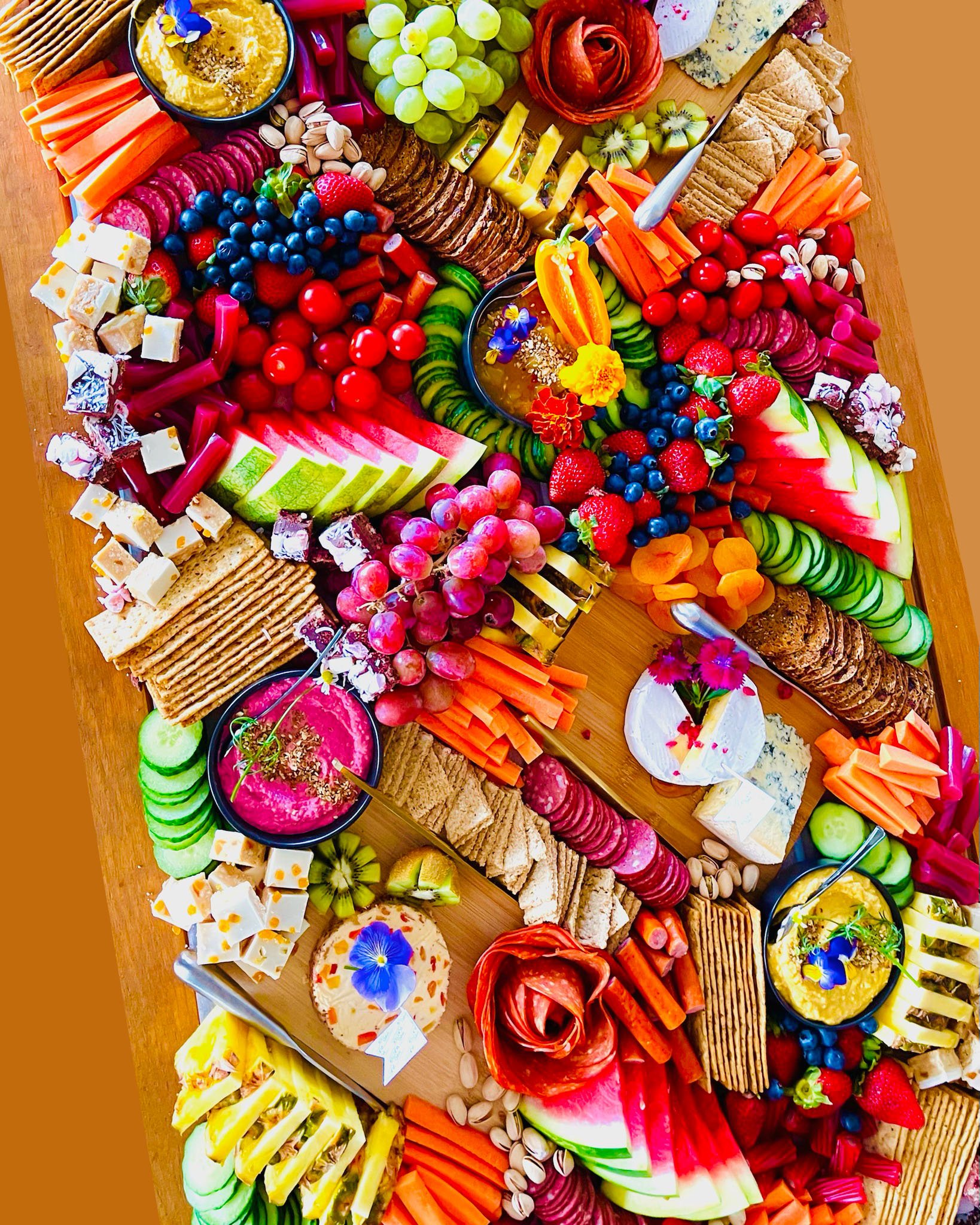 Colorful cheese, fruit, vegetable, and cracker platter with grapes, strawberries, blueberries, sliced cucumbers, carrots, watermelon, cheese cubes, crackers, and dips decorated with edible flowers.