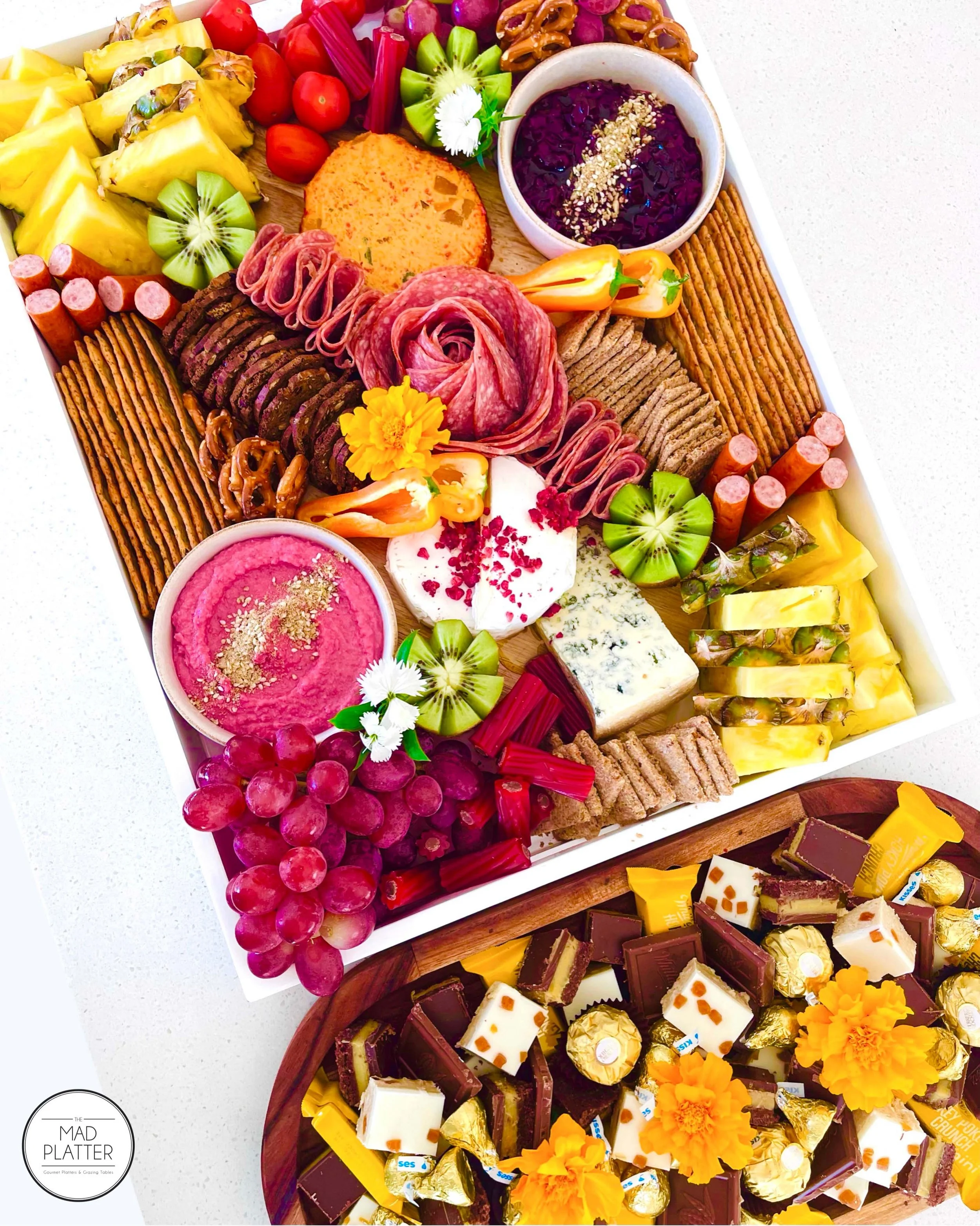 A large decorative tray filled with an assortment of fresh fruit, cheeses, crackers, dips, chocolates, and flowers, arranged for a celebration or gathering.