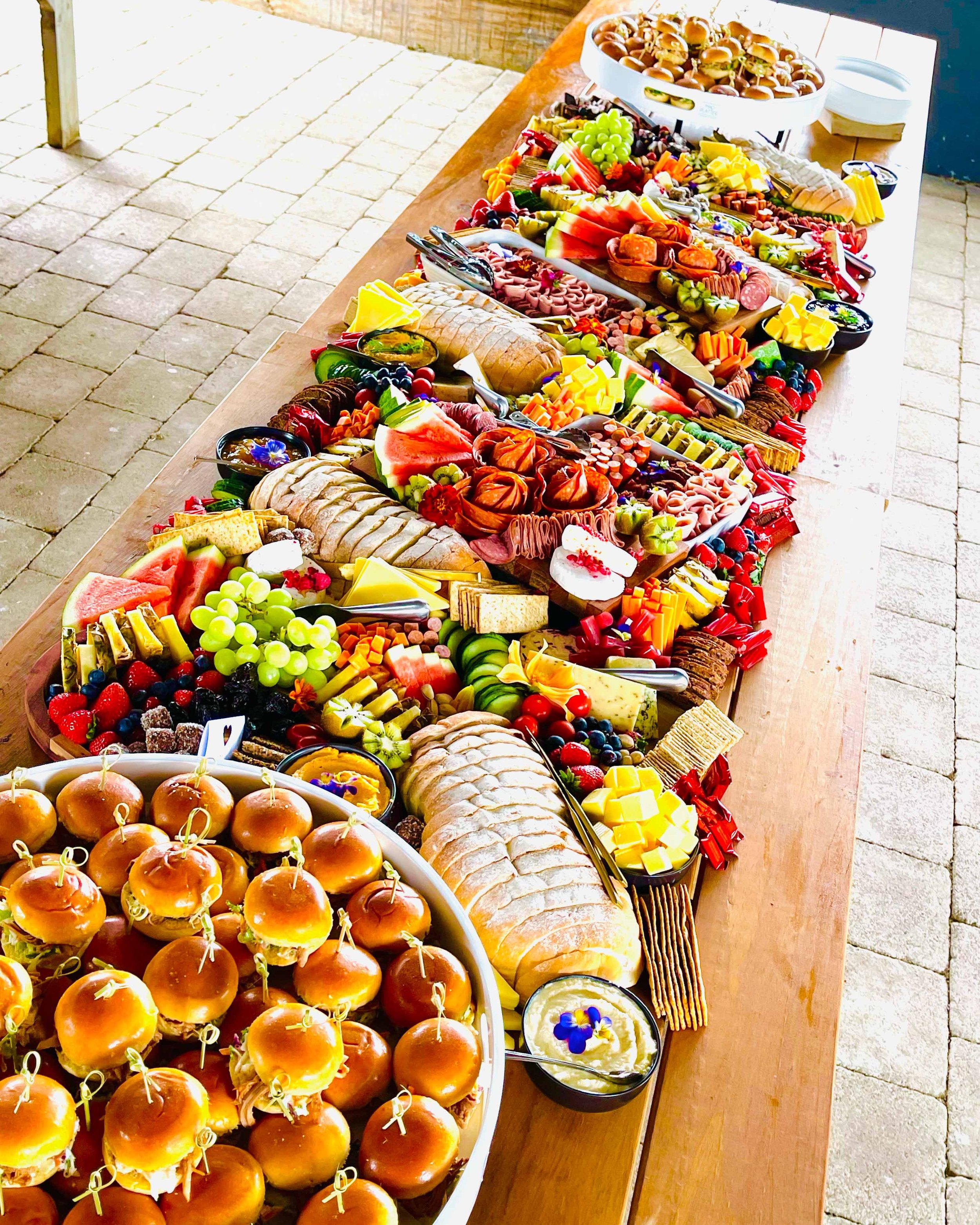 A long wooden table filled with various foods including mini sliders, grapes, watermelon, cheese, and fruits, with bowls of dips and condiments, set on an outdoor stone patio.