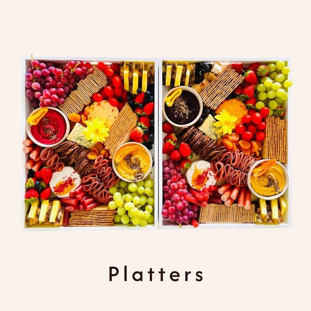 Two white boxes filled with assorted crackers, fruits, cheese, and dips, labeled as "Platters."
