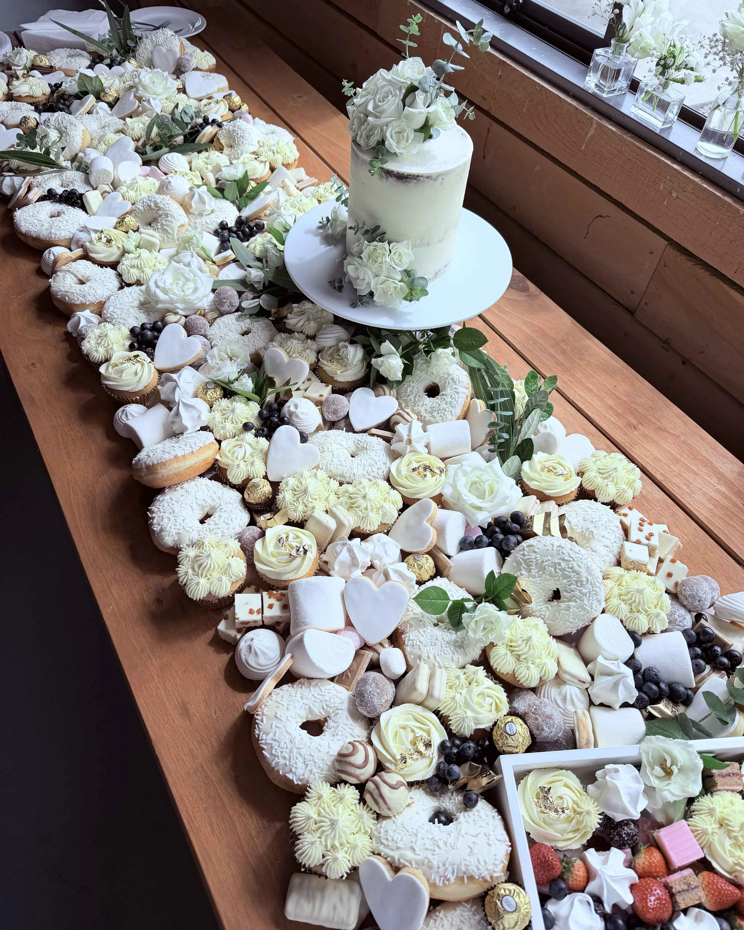 A large assortment of decorated cookies topped with white, yellow, and pastel icing, and some with sprinkles, chocolate, and fruit decorations, arranged on a long wooden table with a floral centerpiece and a tiered cake decorated with white roses.