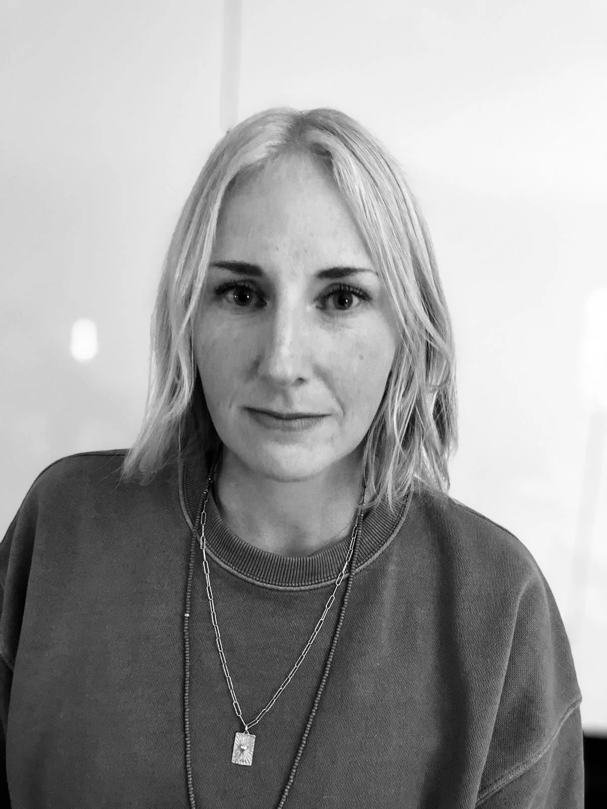 Black and white photo of a woman with shoulder-length hair, wearing a casual top and layered necklaces, looking directly at the camera.