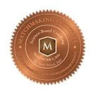 A bronze-colored seal or badge with a central shield containing the letter 'M', surrounded by text and decorative elements.