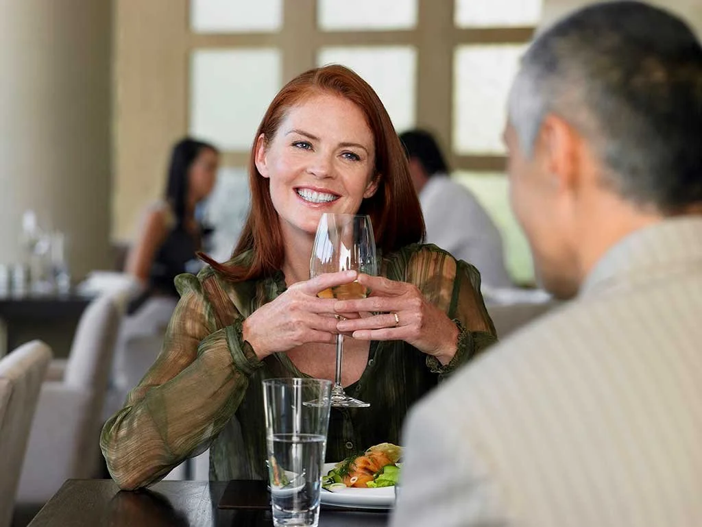 A woman with red hair smiling and holding a glass of white wine while sitting at a restaurant table across from a man. The table has a water glass and a plate of food.