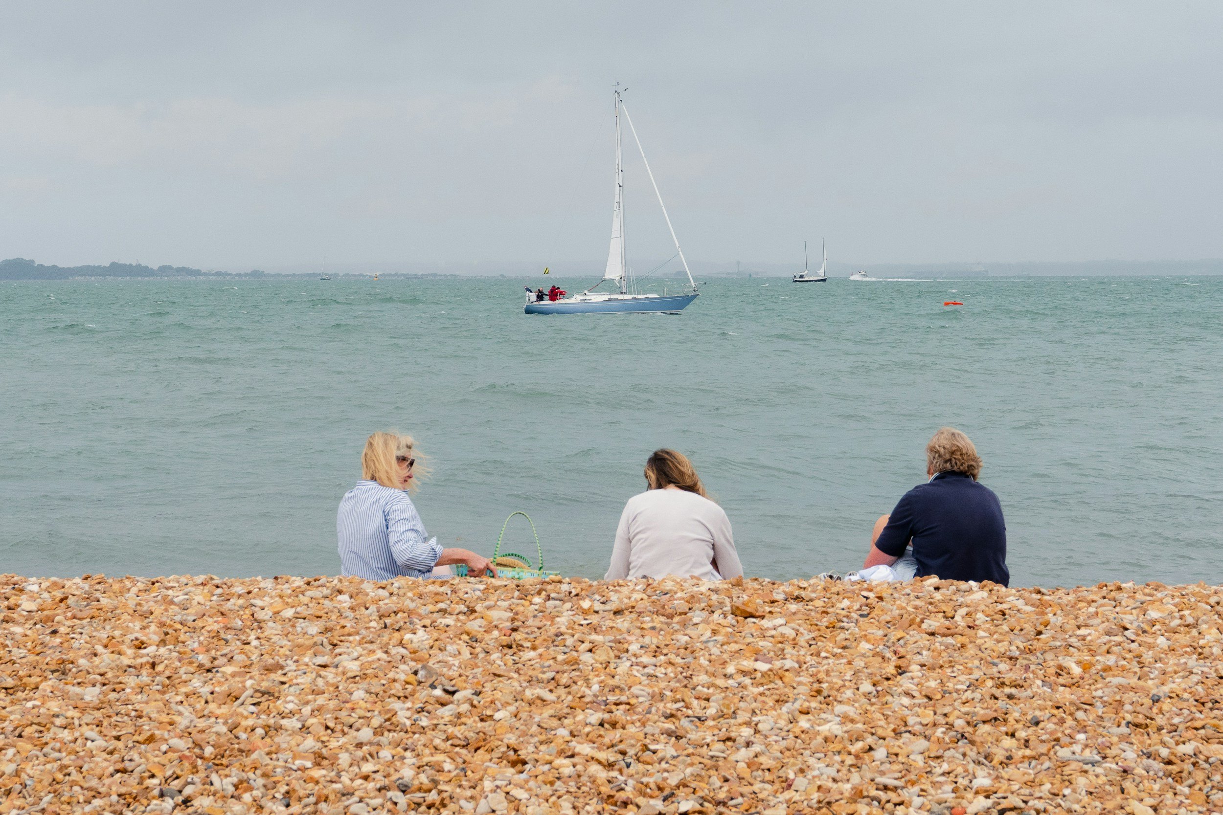 People sitting on a pebble beach watching sailboats on the water beneath a cloudy sky.