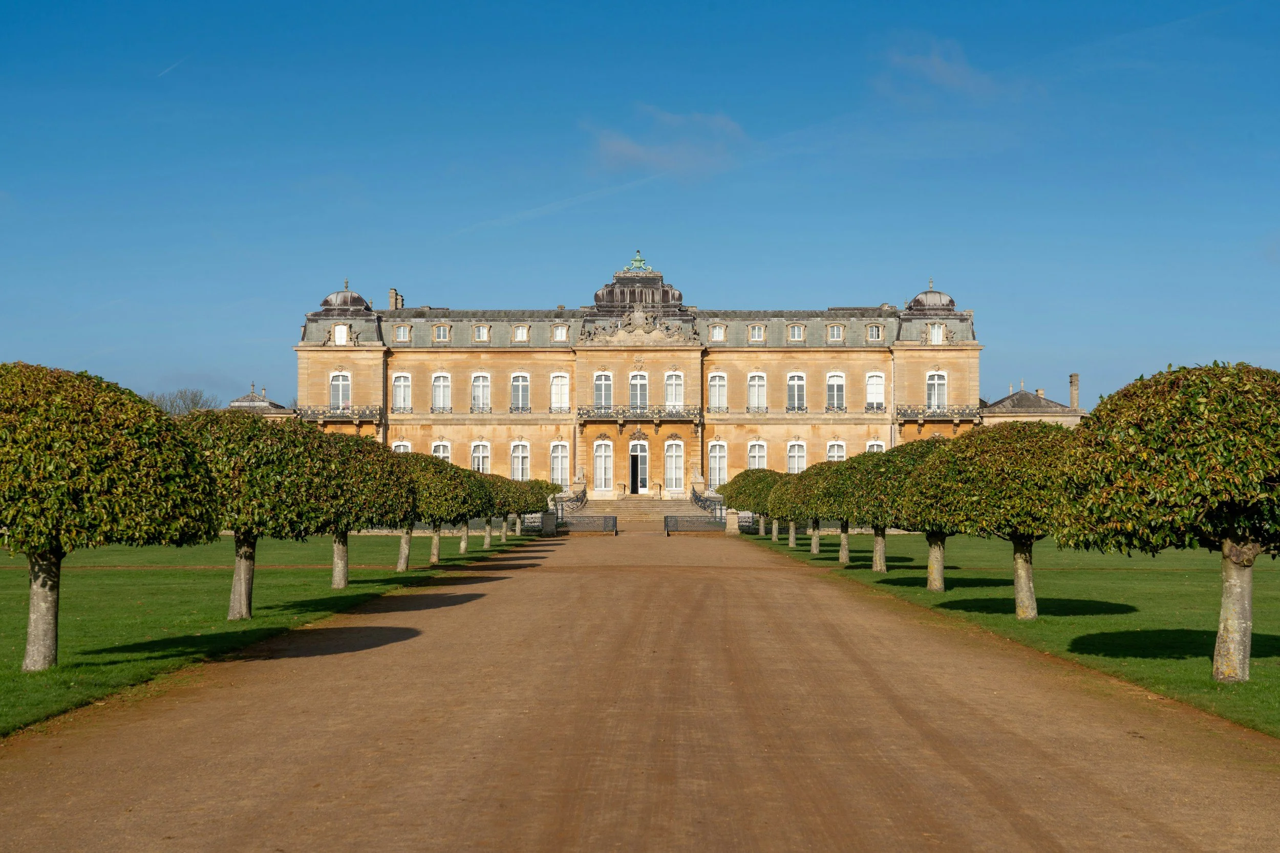 A grand historic mansion with a symmetrical facade, surrounded by neatly trimmed trees and a wide pathway leading up to it under a clear blue sky.