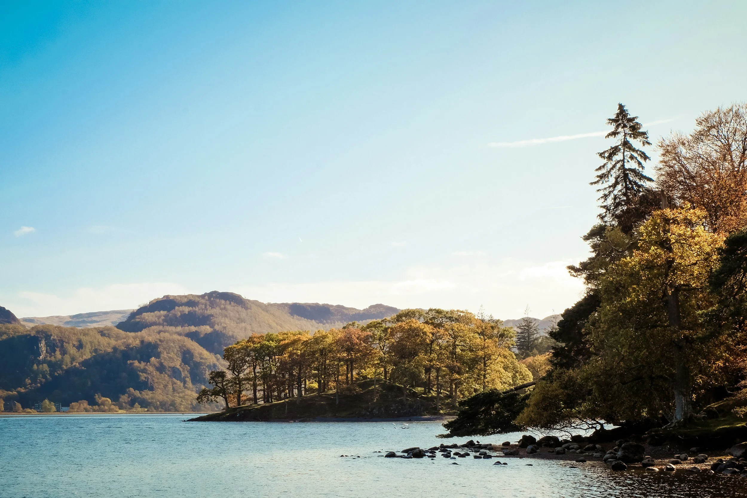Scenic view of a lake with calm waters, surrounded by hills and dense trees, some with fall foliage, under a clear blue sky.