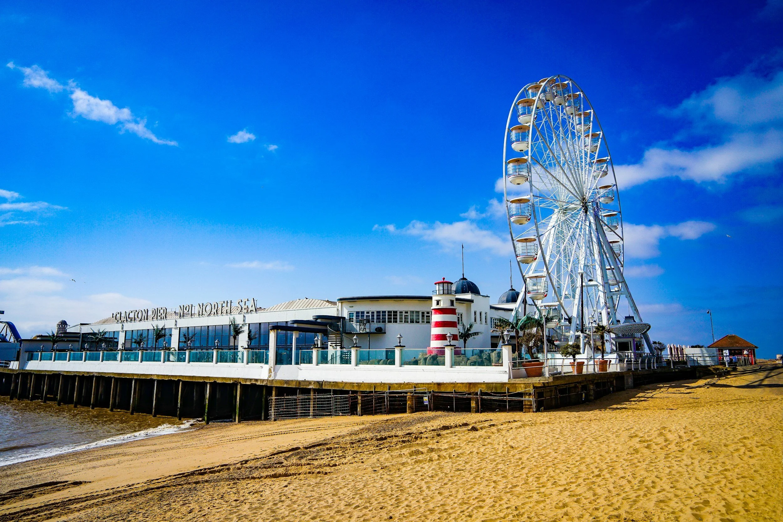 A full view of the Santa Monica Pier with a large Ferris wheel and buildings against a bright blue sky, with sandy beach and water in the foreground.